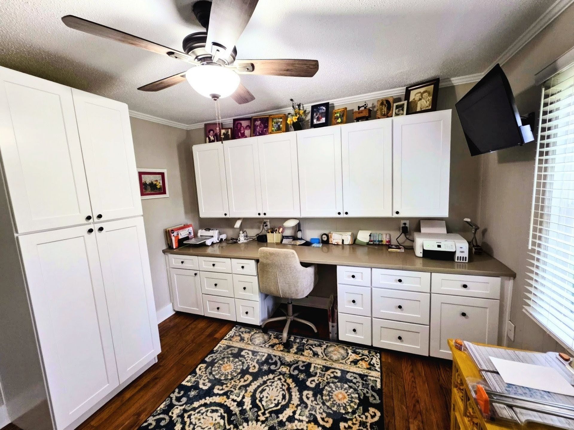 Home office with white cabinets, built-in desk, rolling chair, printer, ceiling fan, and a rug on wood flooring.