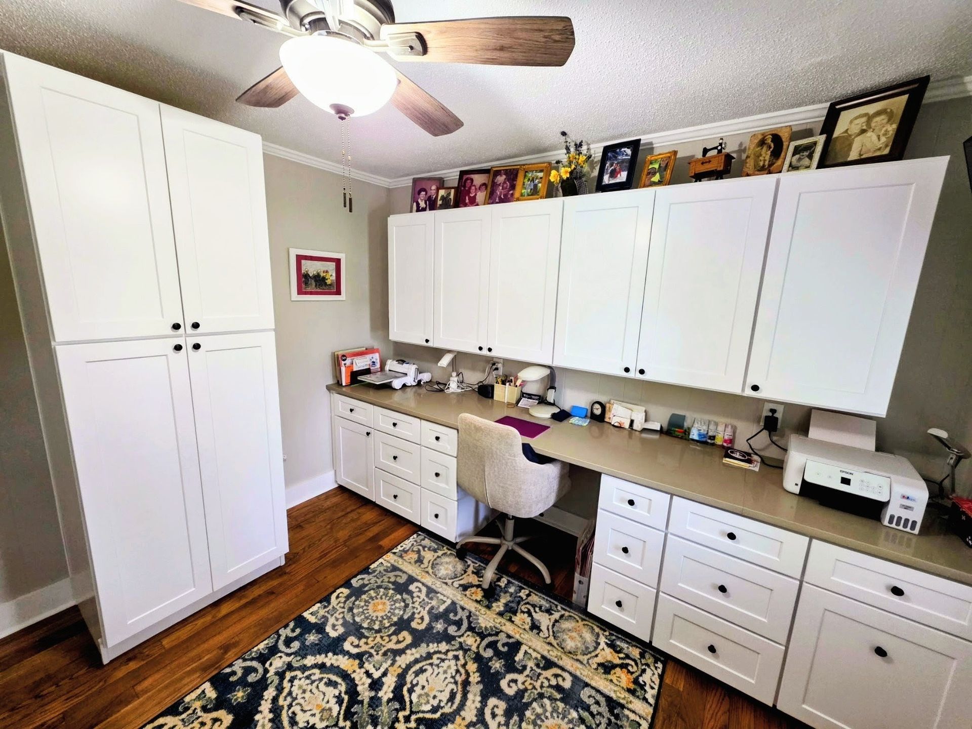 A home office with white cabinetry, a desk, a printer, and a patterned rug on hardwood floors under a ceiling fan.