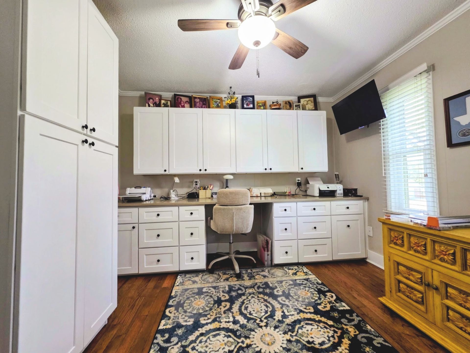 A home office with white built-in cabinets, a desk chair, a patterned rug, and a yellow wooden cabinet on wood floors.