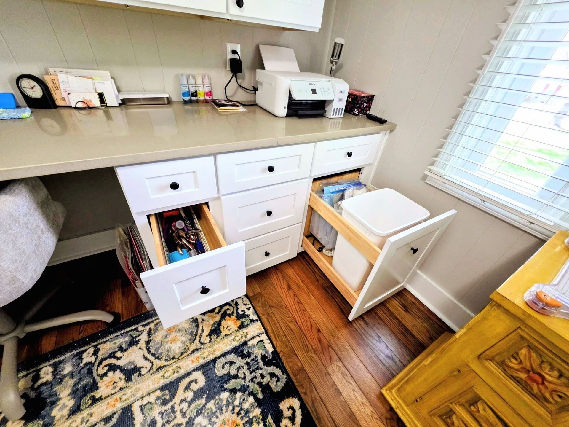 A white desk with an open drawer and a pull-out trash bin unit next to a window in a home office setting.