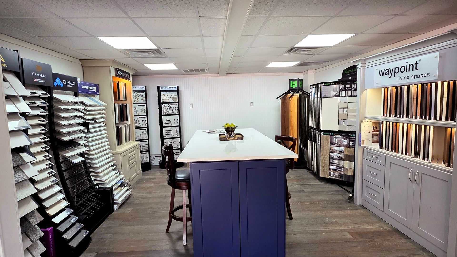 Flooring store showroom with sample displays, a central island, and shelves of carpet and tile samples.
