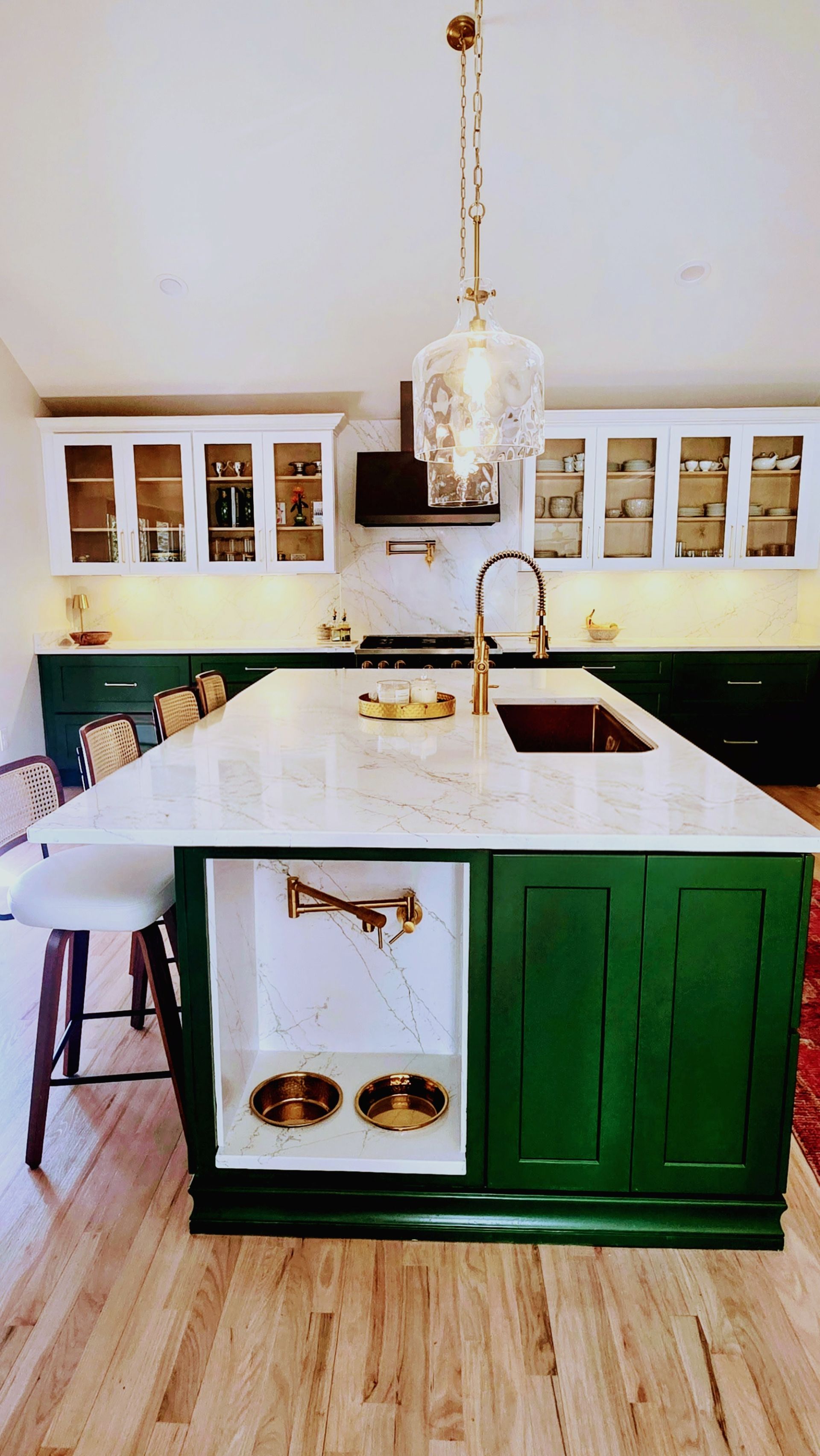 Green kitchen island with pet bowls, gold faucet, pendant light, white marble countertop, and white and green cabinets.