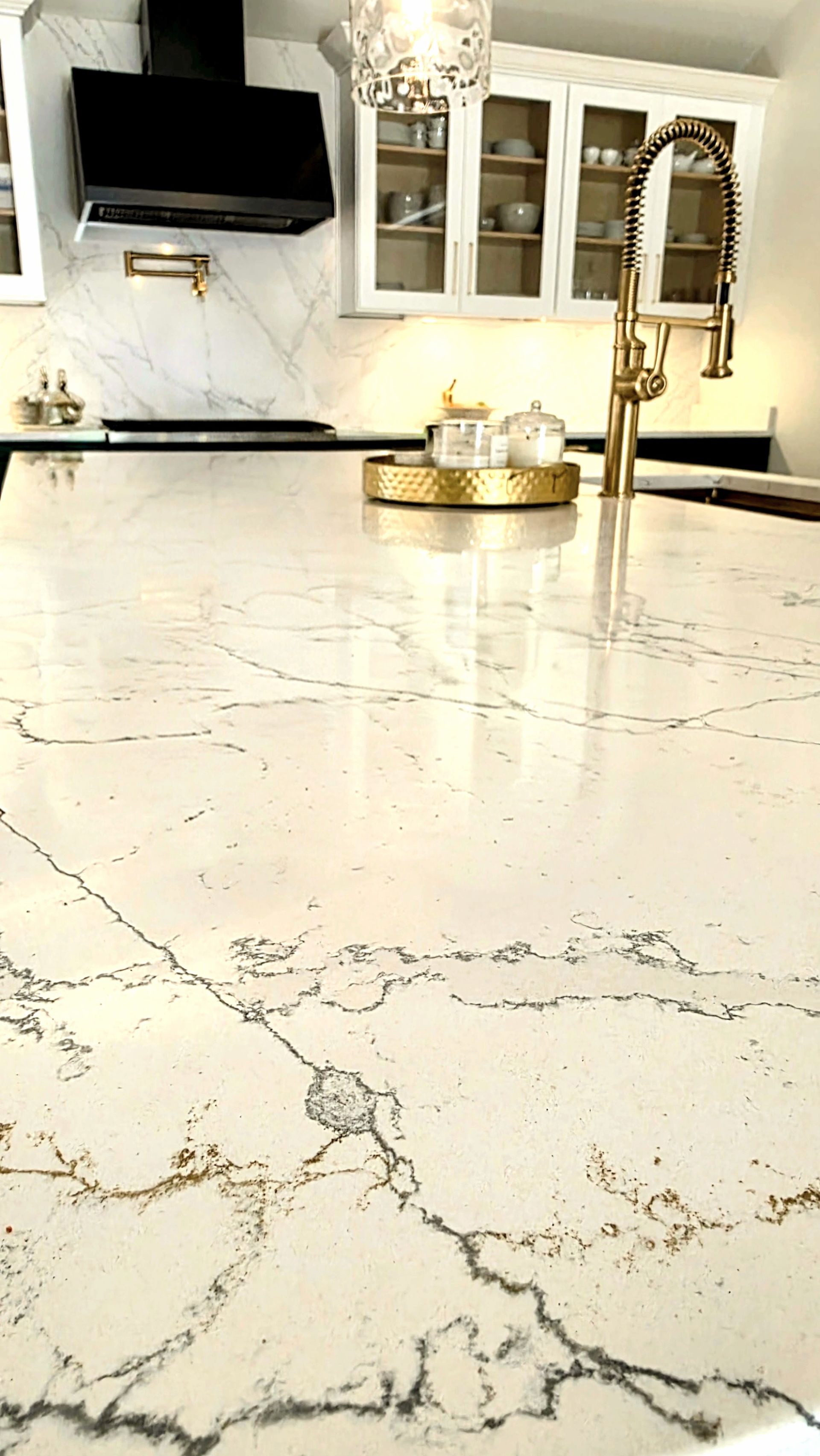 Close-up of a kitchen island with a white countertop featuring gray and gold veining, gold faucet, and a black range hood.