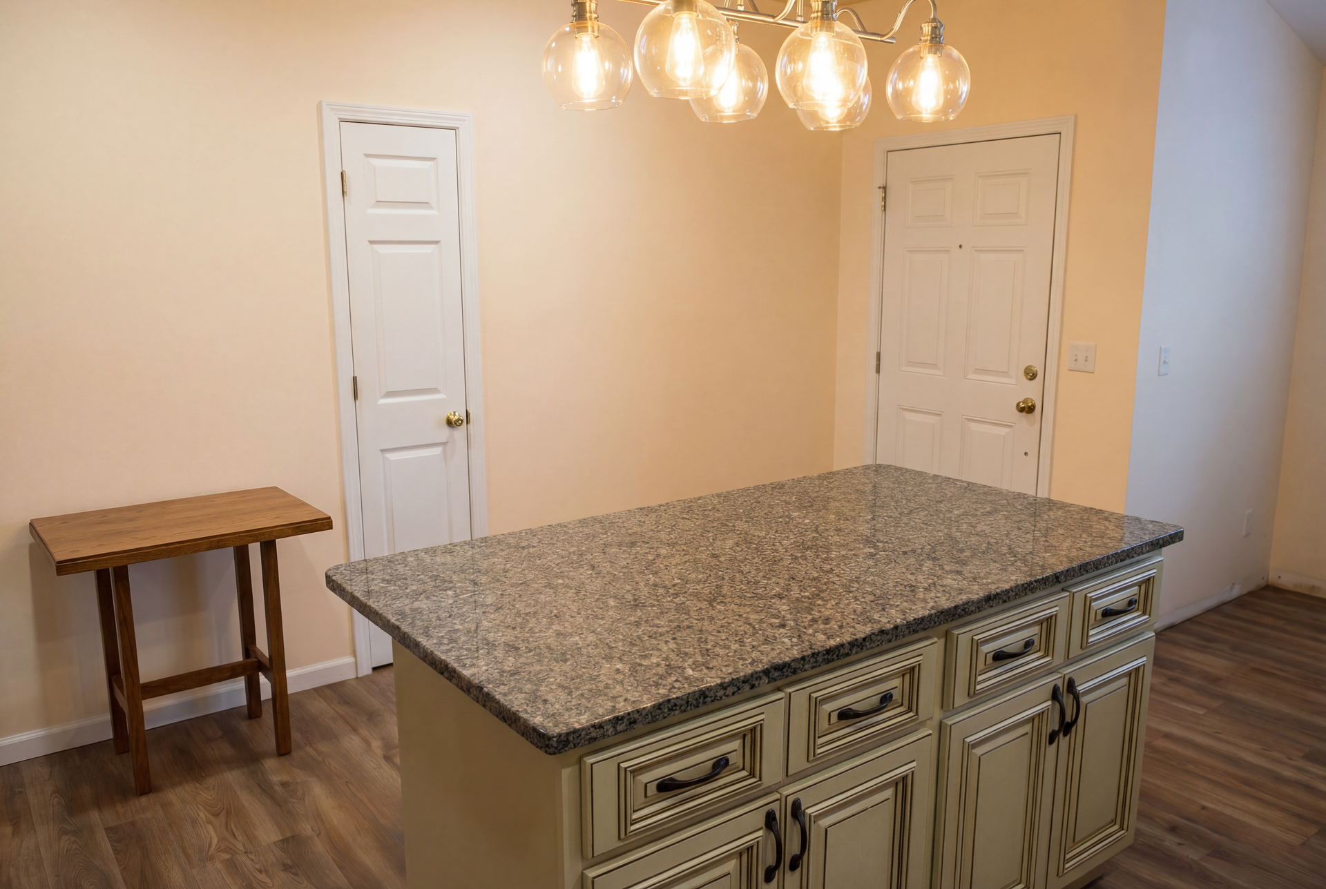 Kitchen with island, doors, and a small table. Granite countertop, light fixture, beige walls, and wood floor.