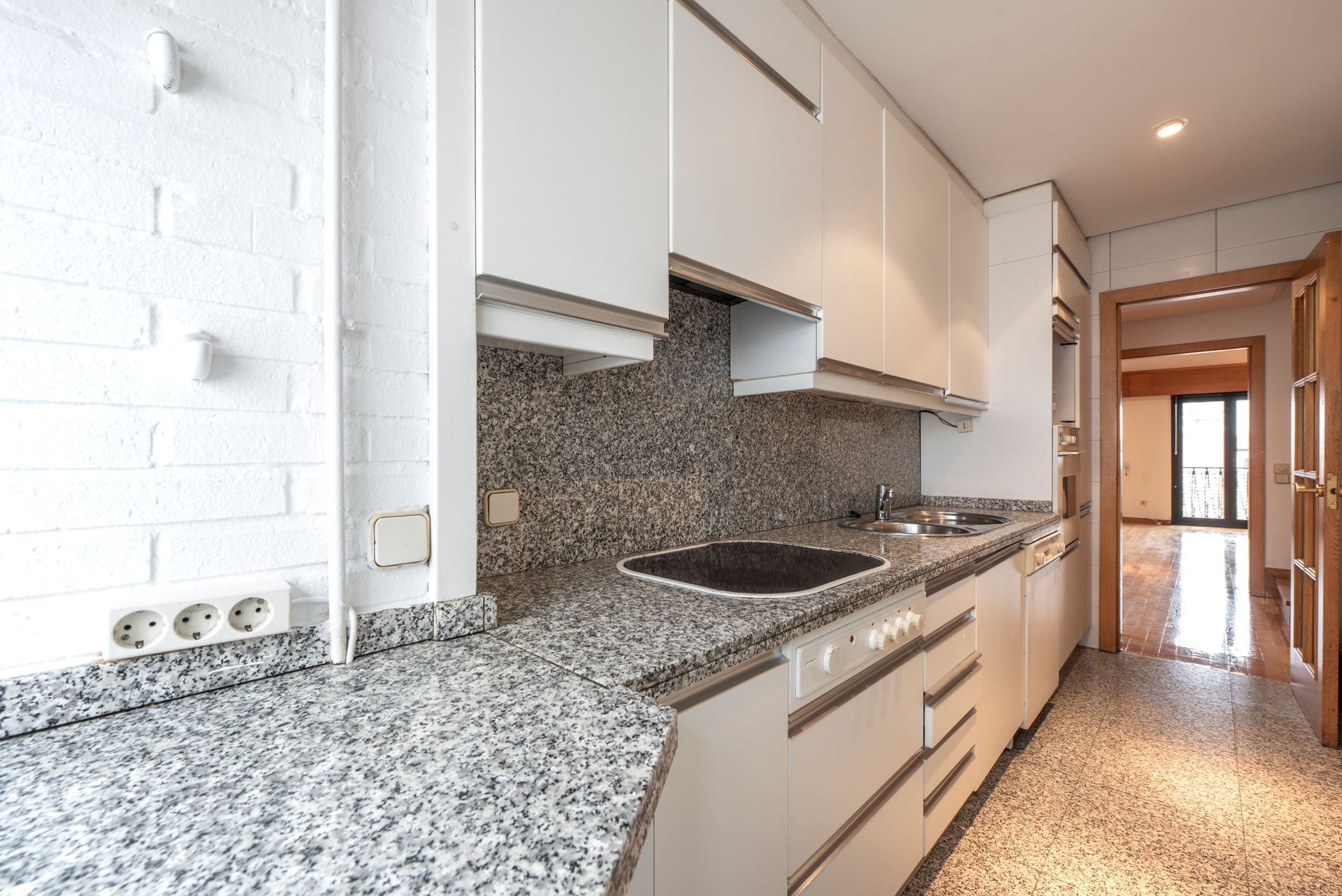 Modern white kitchen with granite countertops and flooring, leading to a hallway.