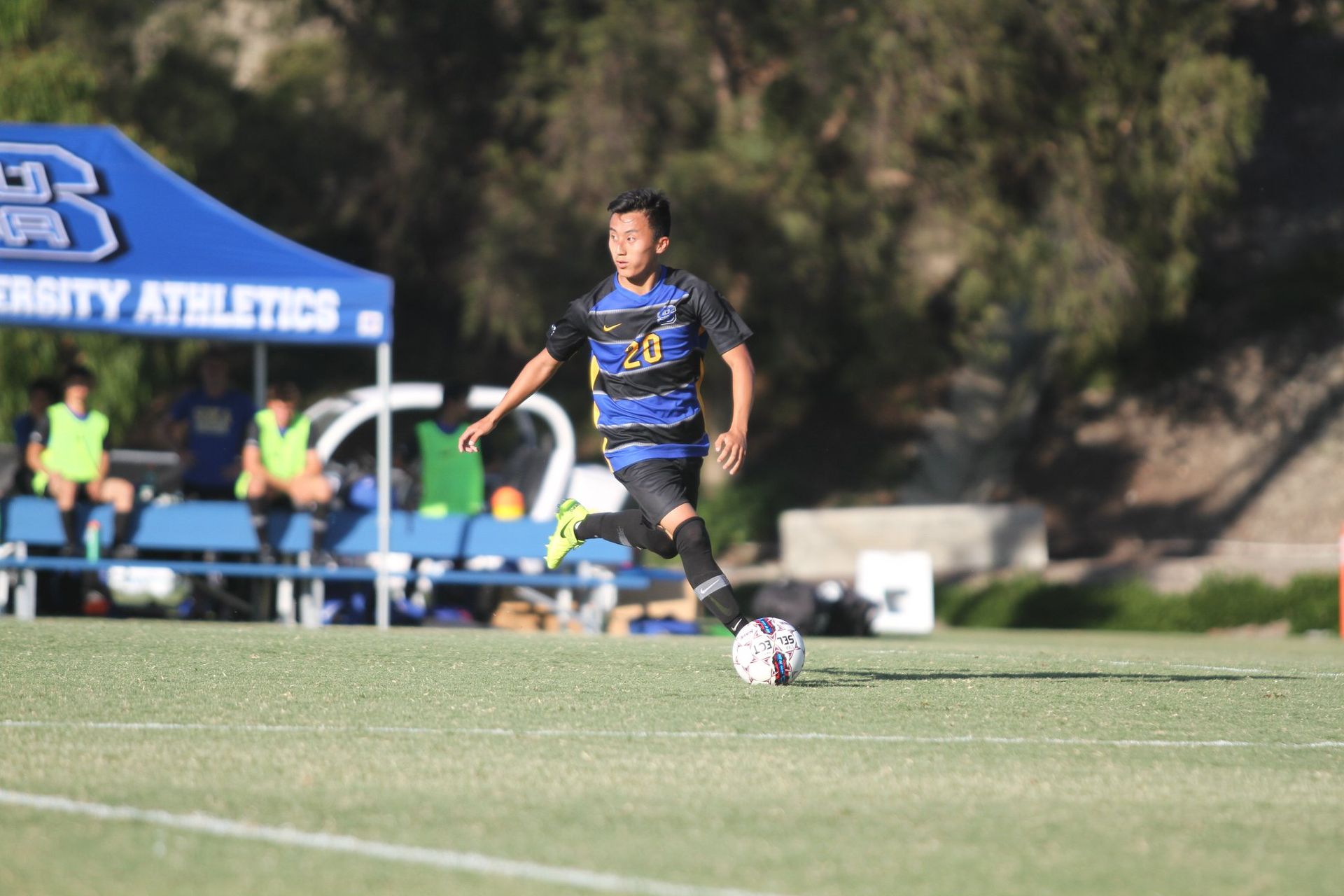 A man is kicking a soccer ball on a field with a university athletics tent in the background.