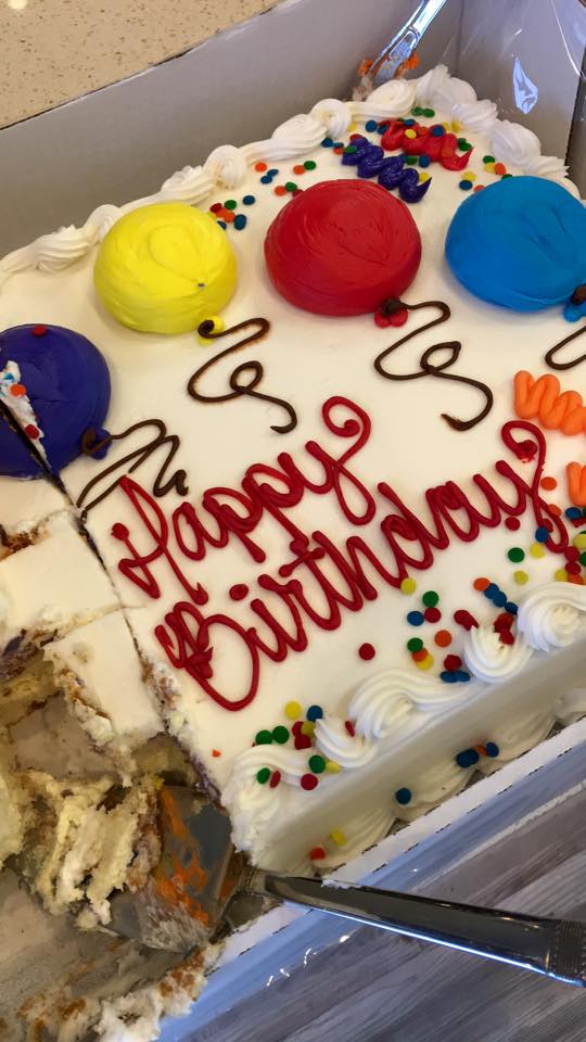 A birthday cake with balloons and sprinkles has a slice taken out of it
