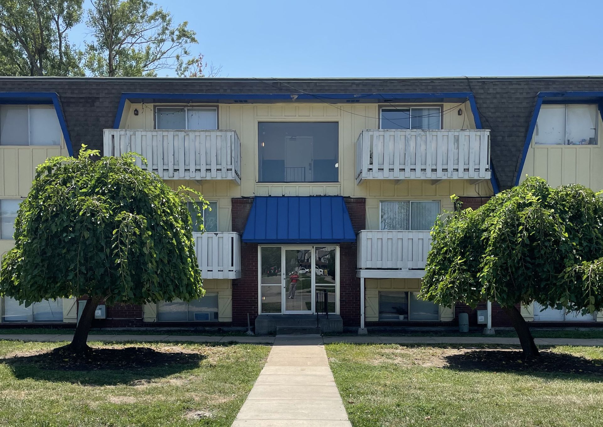 Apartment building with yellow walls, blue accents, balconies, and weeping trees.
