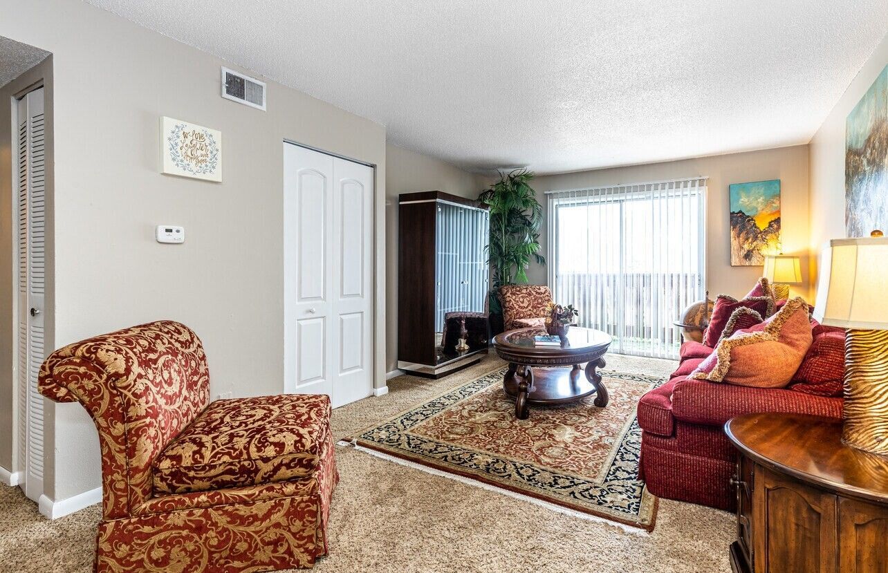 Living room with patterned chair and sofa, rug, sliding glass door, and a brown table with a lamp.