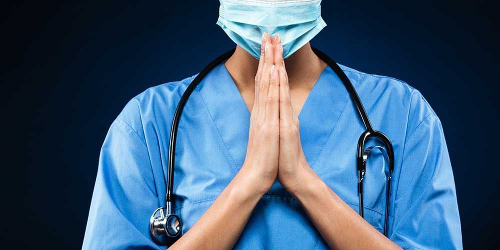 A doctor wearing a mask and a stethoscope is praying.