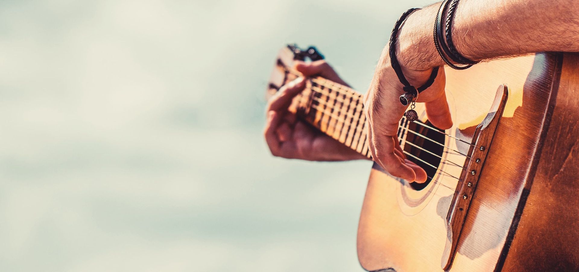 A person's hands playing an acoustic guitar outdoors, sky in background.