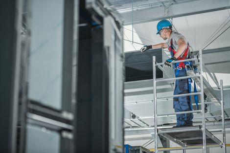 A man is standing on a scaffolding in a building examining duct work.