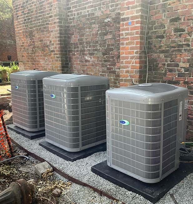 Three air conditioners are sitting on top of gravel in front of a brick building.