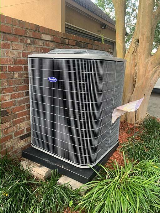 A large air conditioner is sitting in front of a brick wall next to a tree.