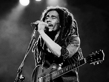 Bob Marley with dreadlocks is singing into a microphone while holding a guitar.