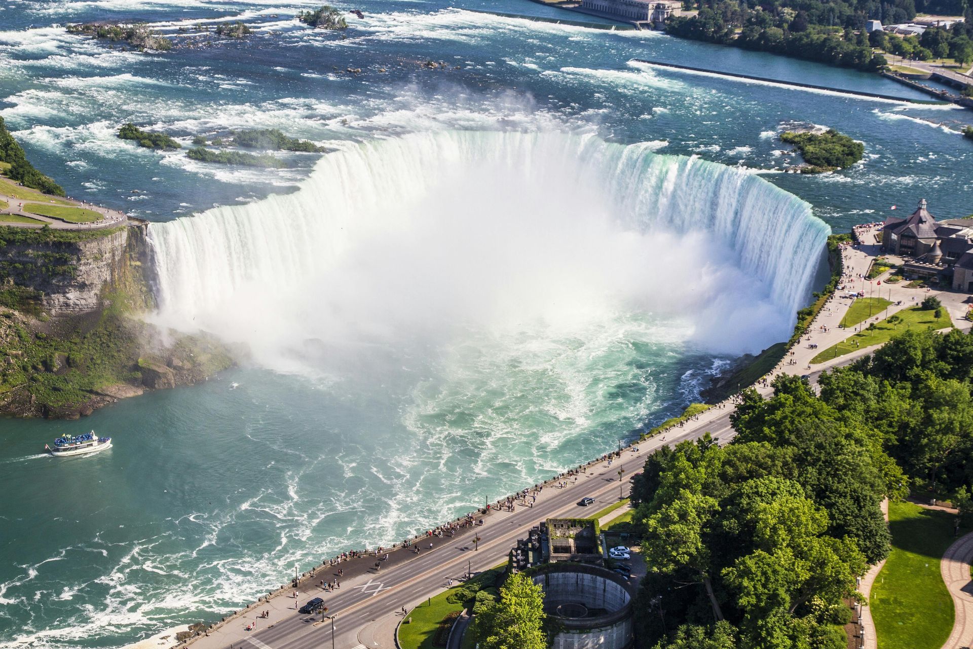 Niagara Falls, viewed from above, with white water cascading over a horseshoe-shaped cliff and a roadway with tourists.