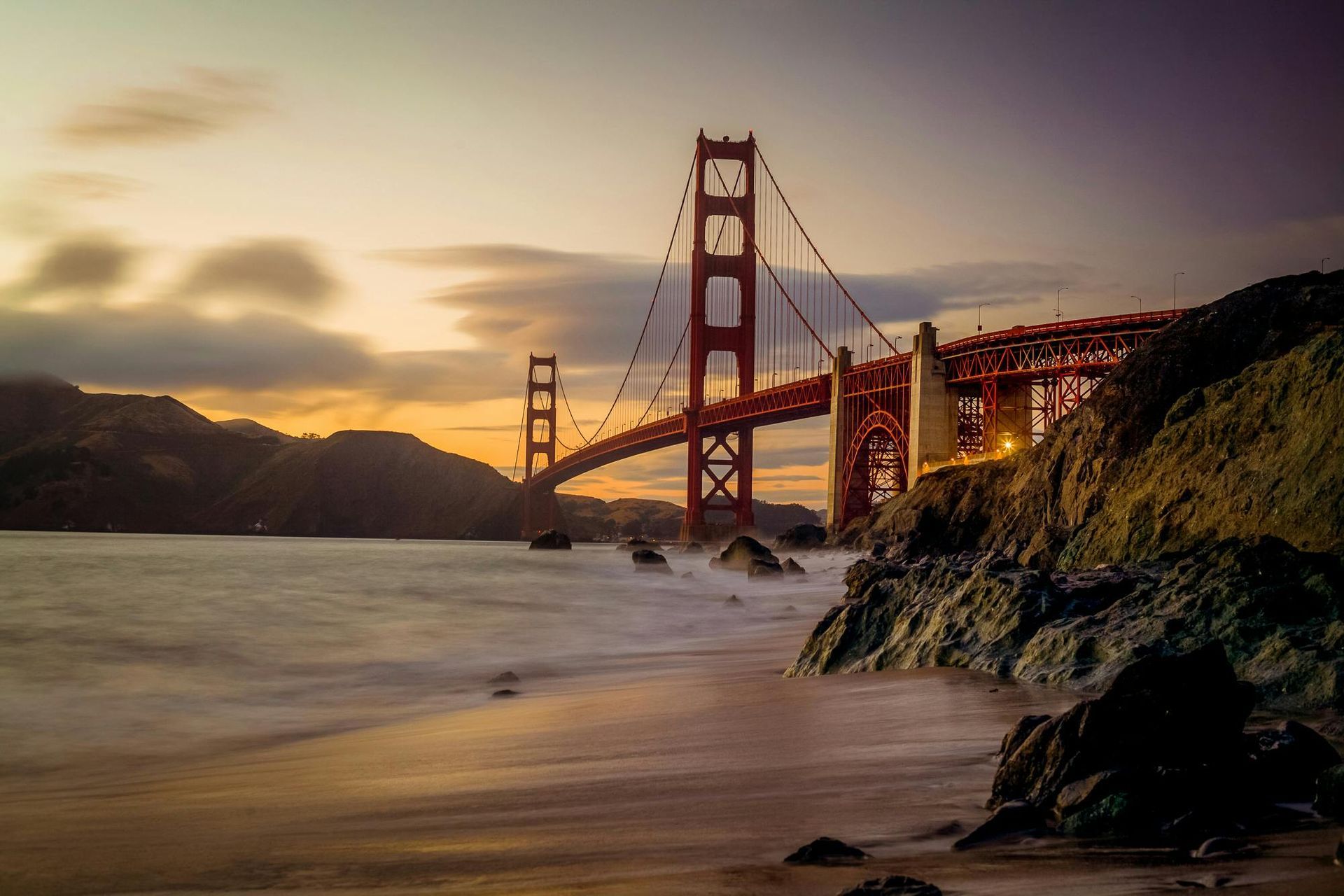 The golden gate bridge is visible from the beach at sunset.