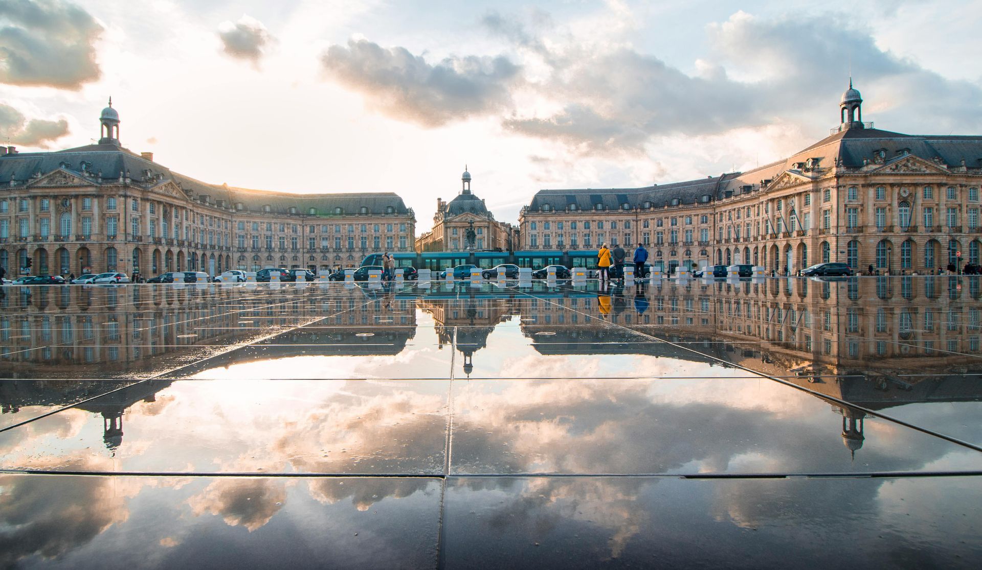 A grand European square with symmetrical buildings reflected in a flooded plaza under a cloudy sky.