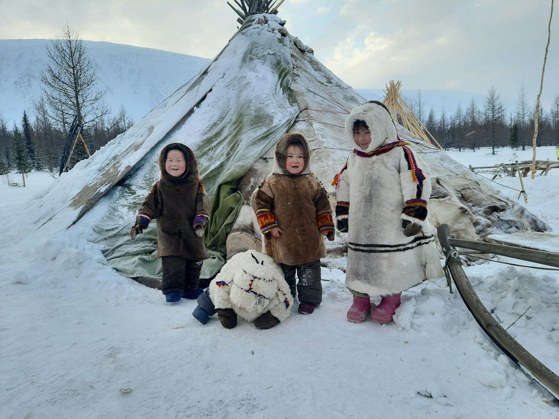 Three children are standing in the snow in front of a teepee.