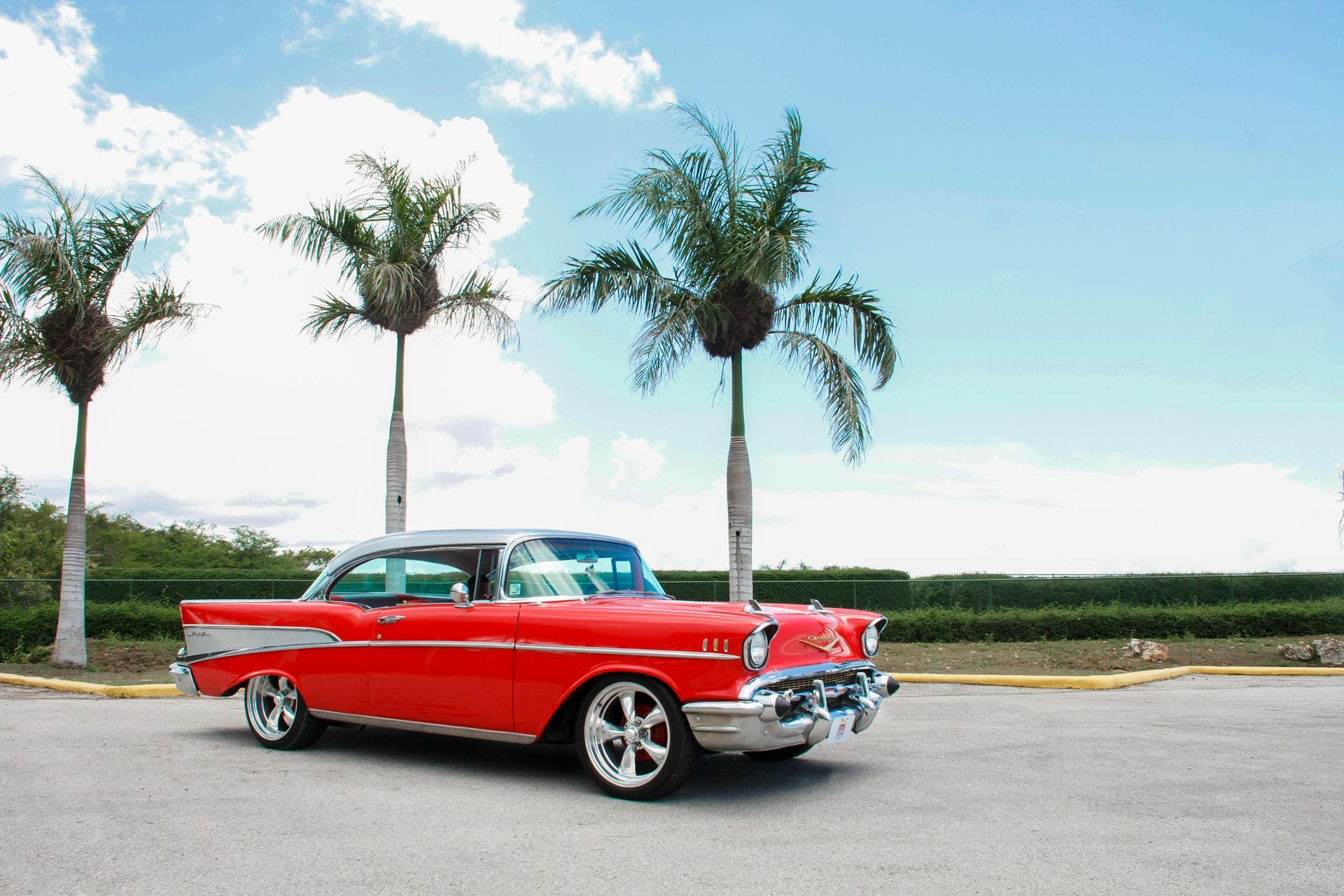 A red car is parked in a parking lot with palm trees in the background