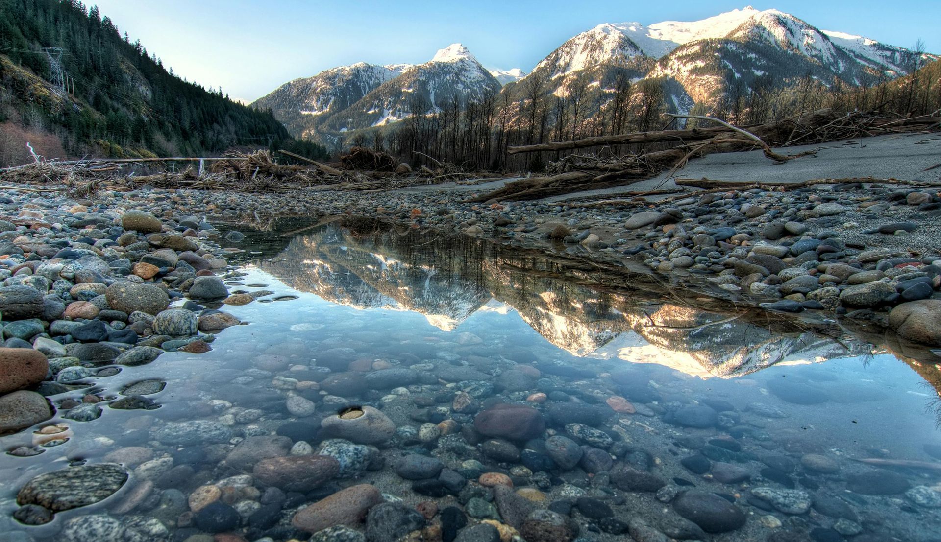 Rocky riverbed reflecting snow-capped mountains under a clear sky; forest on the left.