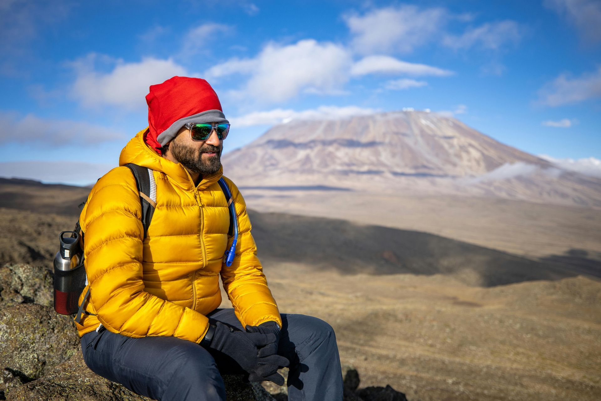 A man in a yellow jacket and red hat is sitting on a rock in front of Kilimanjaro mountain.