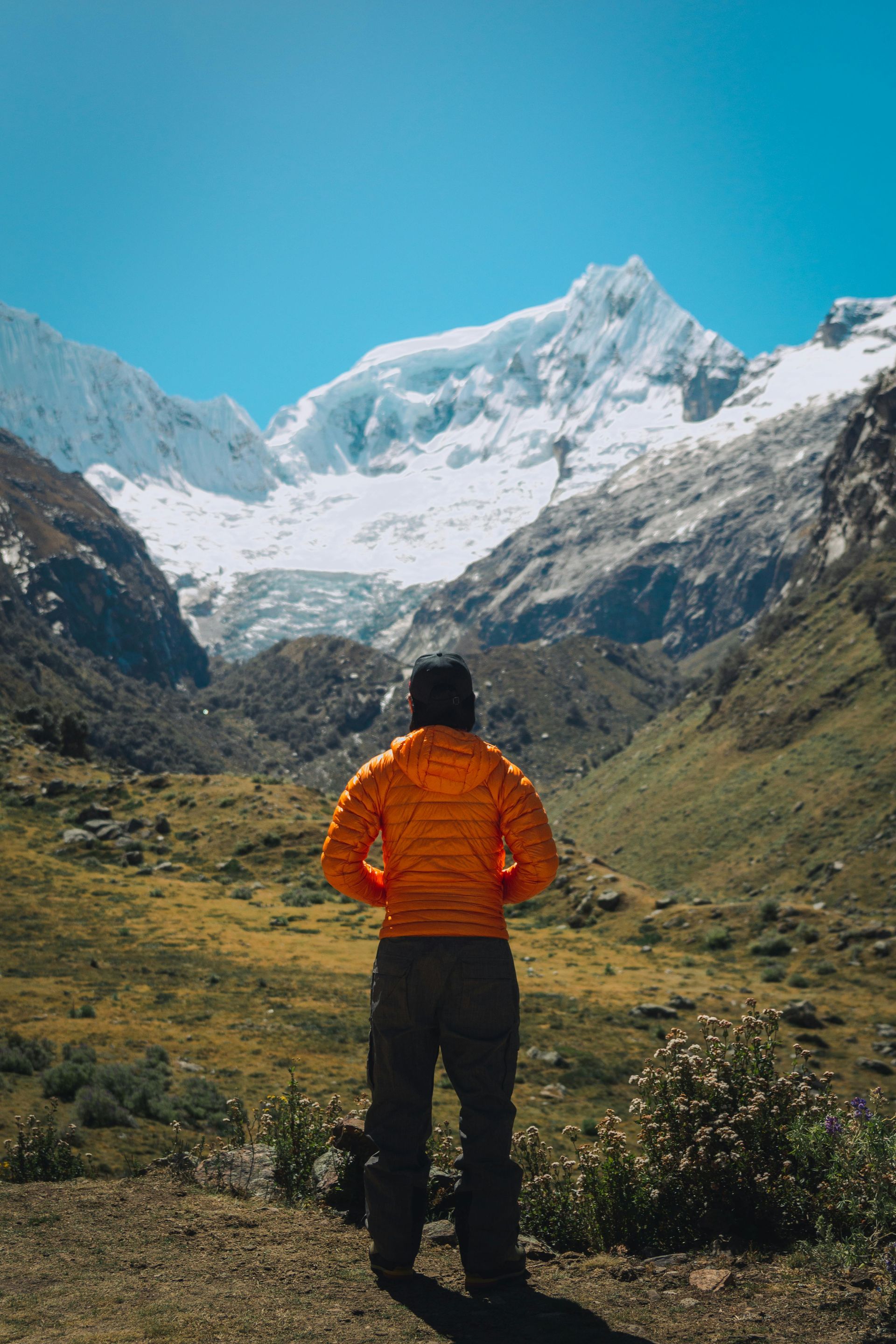 A person in an orange jacket is standing in front of a snowy mountain in the Andes