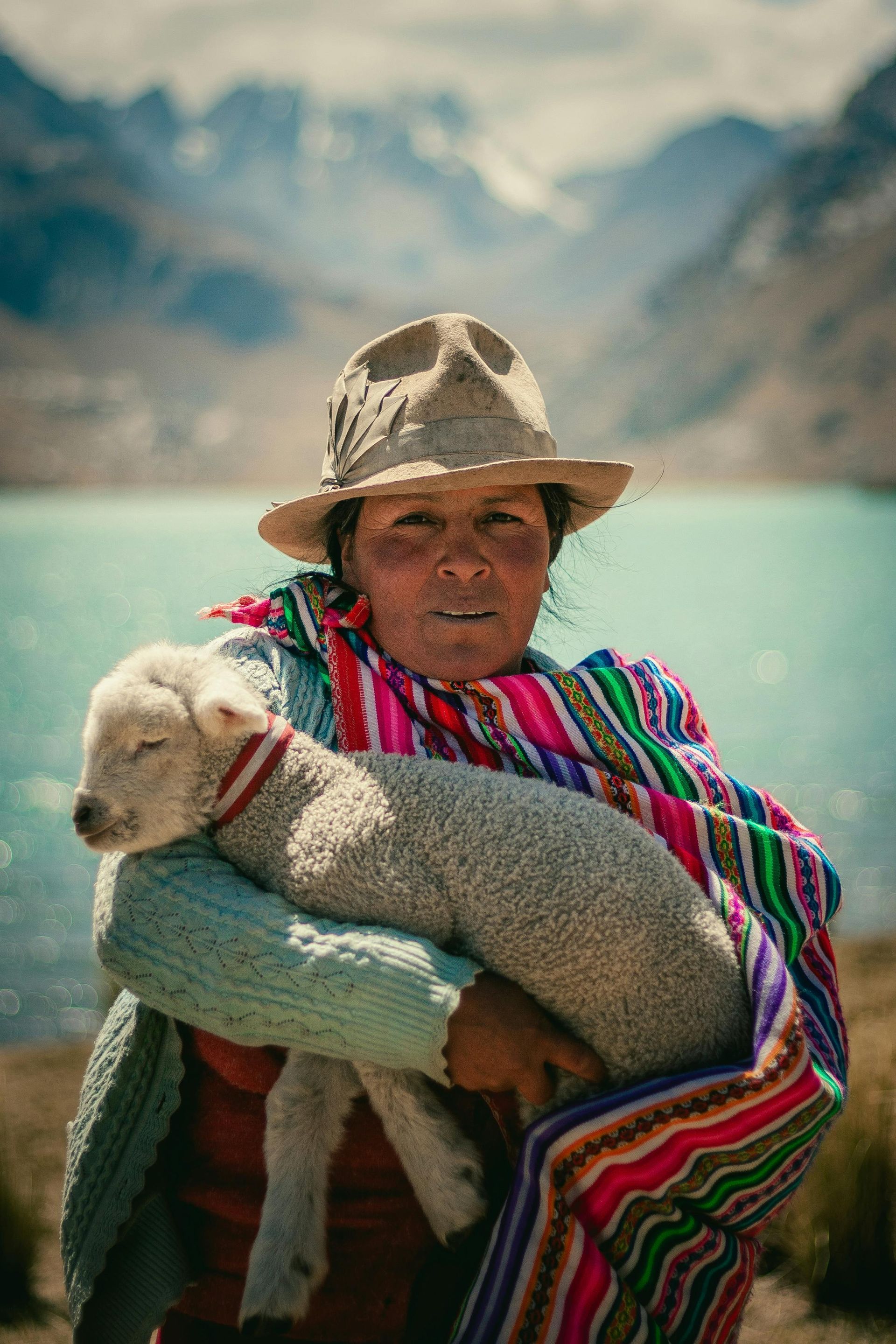 A woman is holding a sheep in her arms in front of a lake in the Andes
