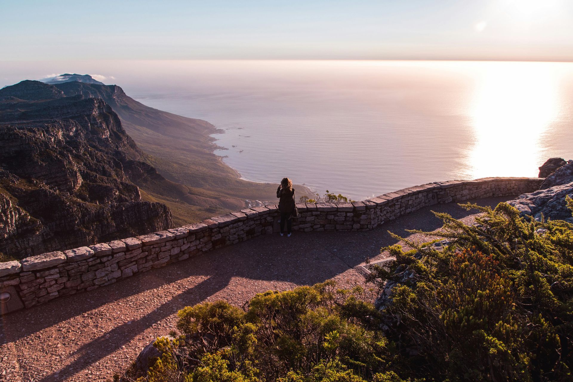 A person is standing on top of a Table Mountain overlooking the ocean.