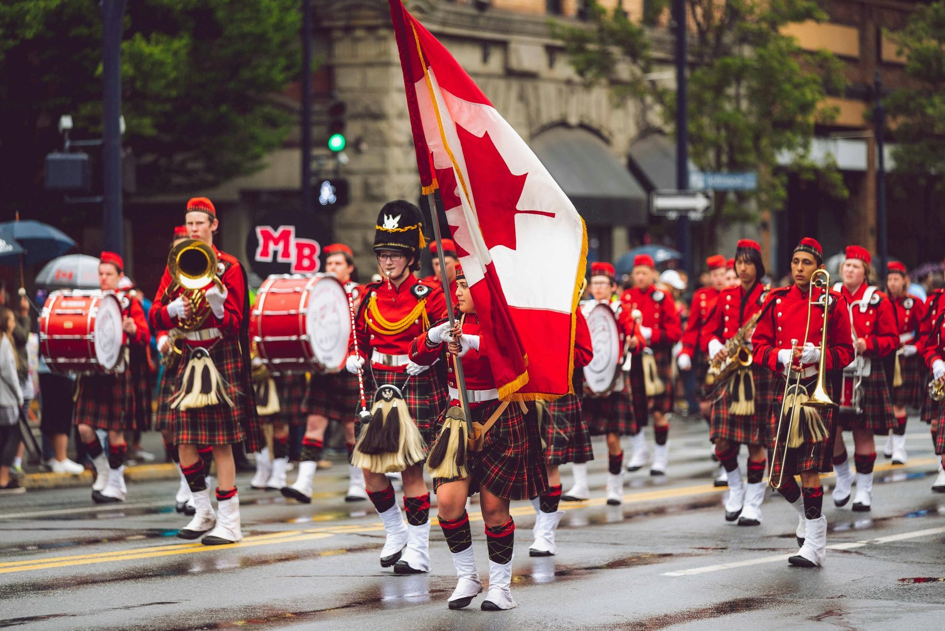 Canadian marching band in red uniforms and kilts parades, holding a Canadian flag, in a city street.