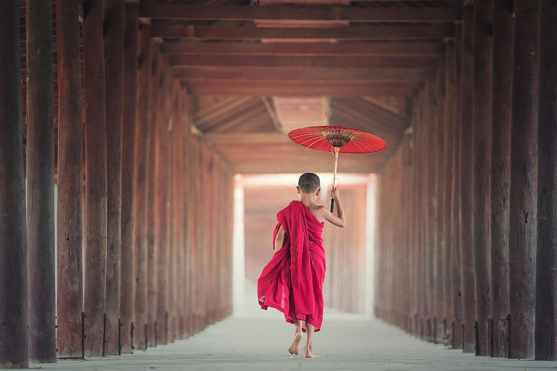 A young monk in a red robe is holding an umbrella in a wooden tunnel.
