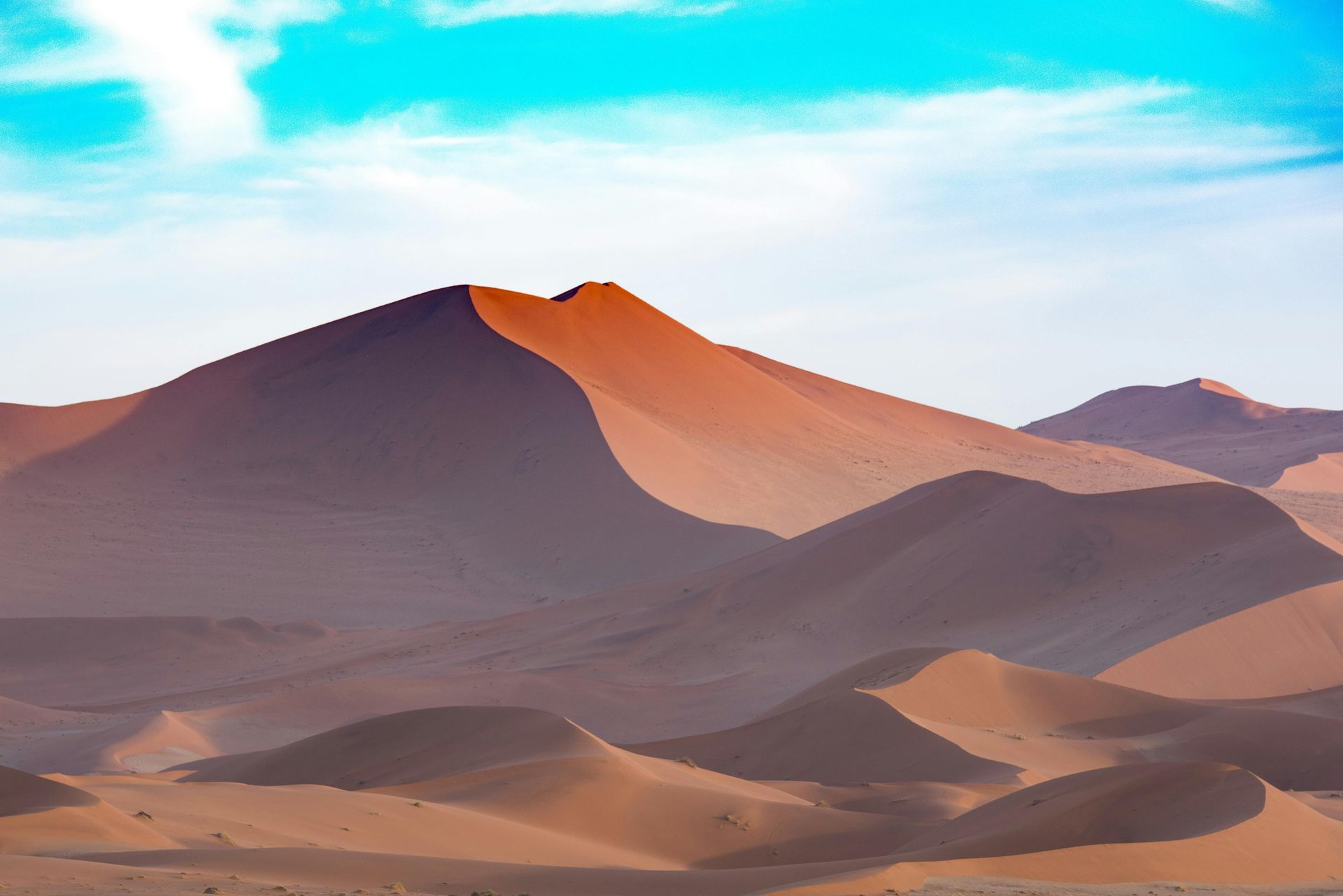 Namib desert landscape with mountains and sand dunes in the foreground and a blue sky in the background.