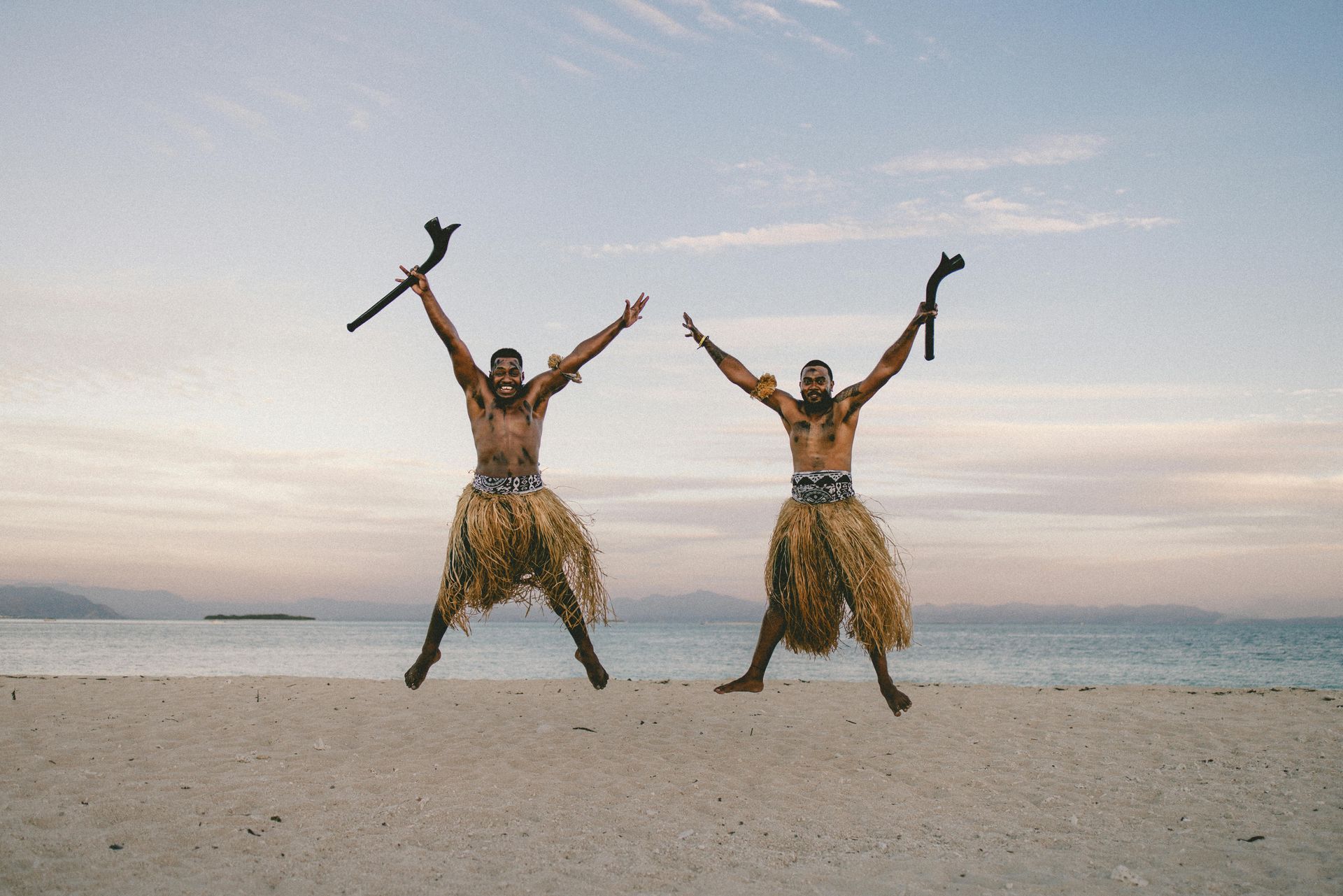 Two men are jumping in the air on a beach with their arms in the air.