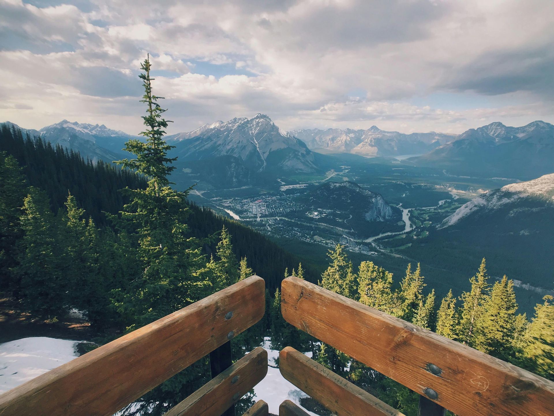 Wooden railing overlooking a mountain valley town. Snow-capped peaks, evergreen trees, and a cloudy sky.