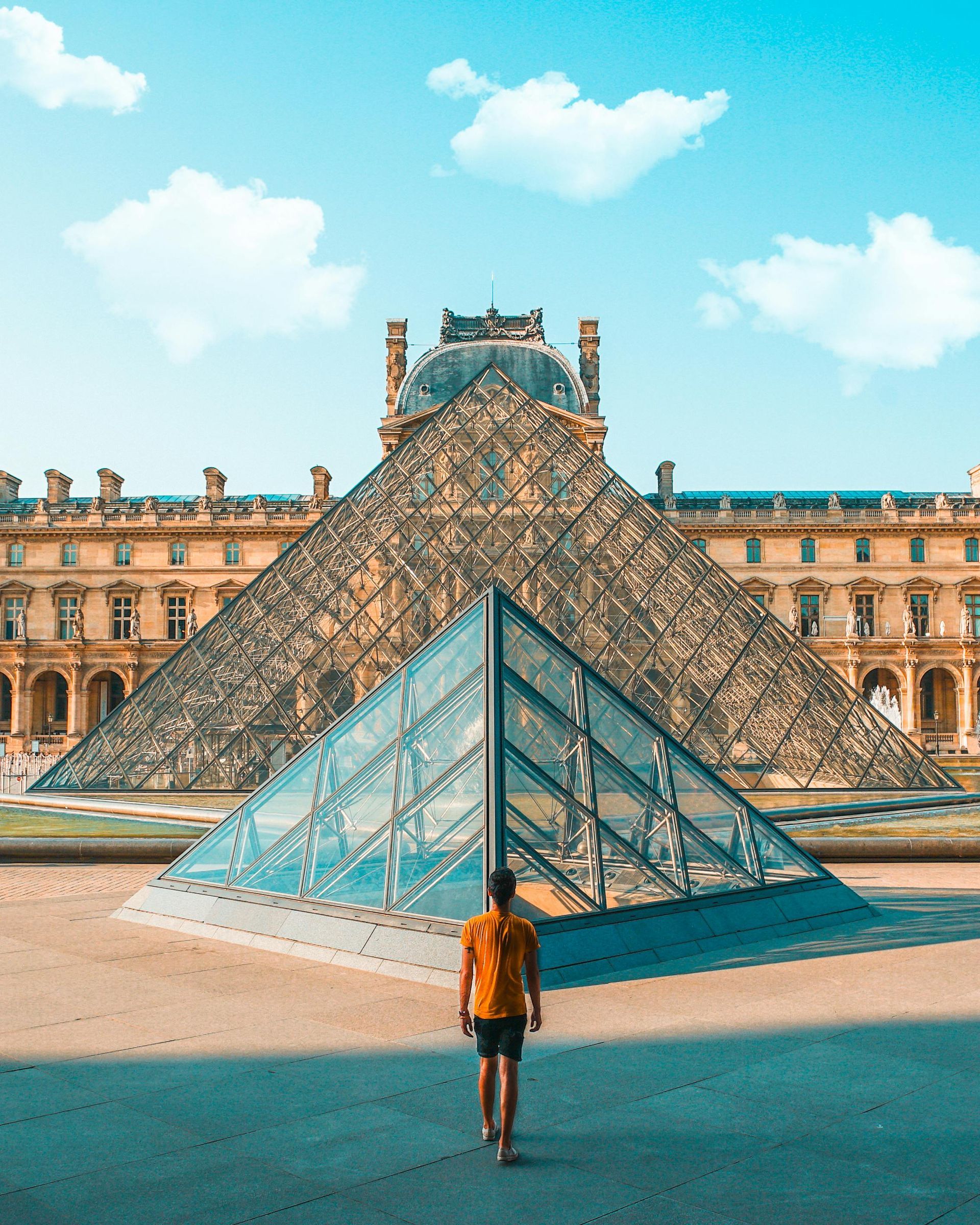 Person walks toward the Louvre Museum's glass pyramid in Paris, France, under a blue sky with fluffy clouds.
