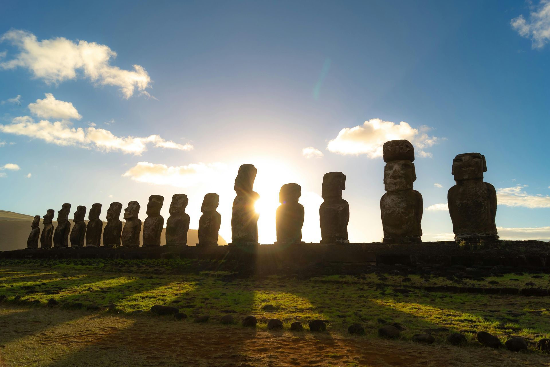 The sun is setting behind a row of Rapa Nui statues in a field.