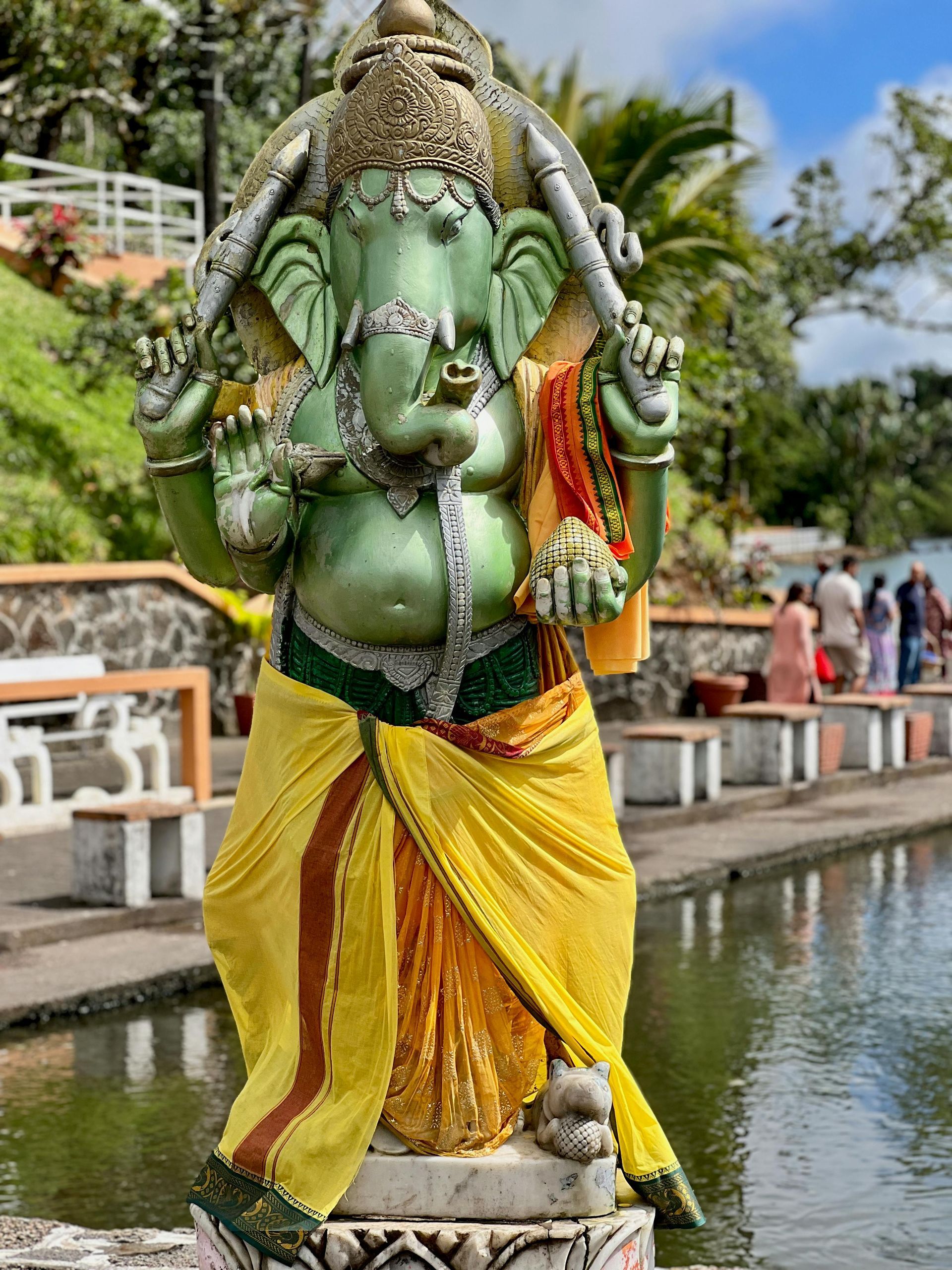 A green and yellow statue of an Indian god deity standing next to a body of water at Grand Bassin