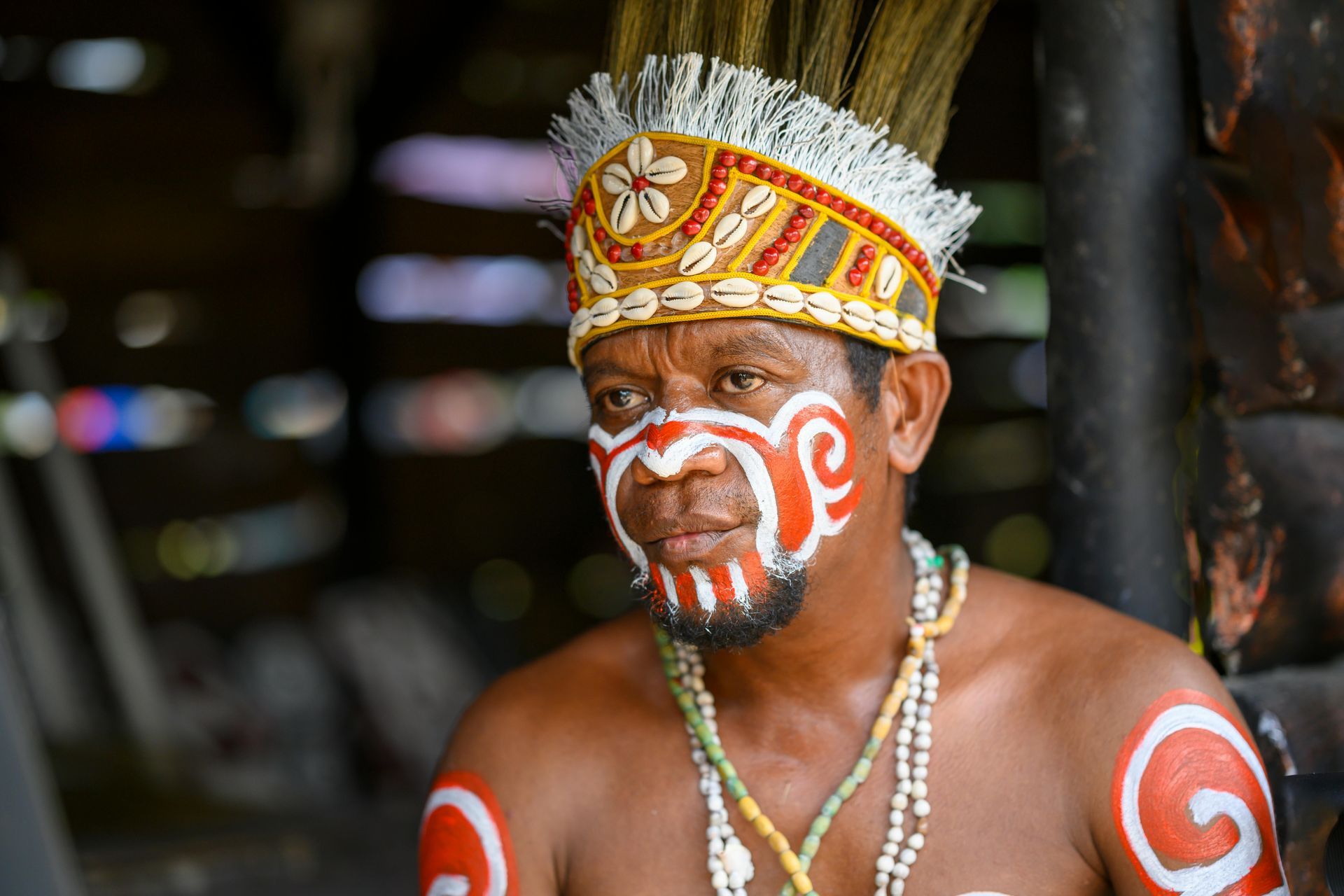 A man with his face painted and a headdress is standing in front of a wooden wall.