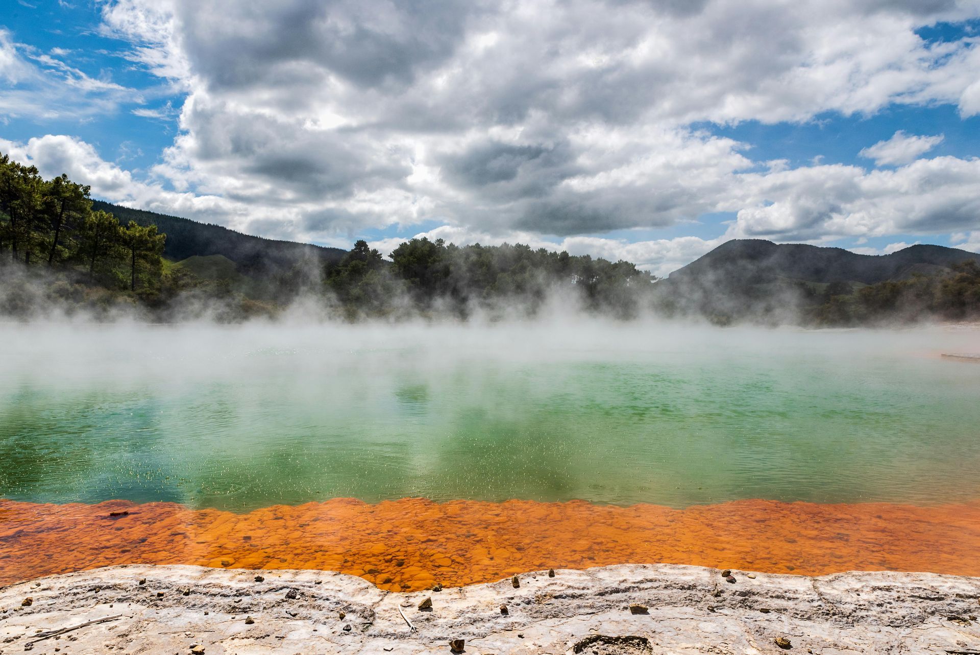 A hot spring with steam coming out of it and mountains in the background in Rotorua
