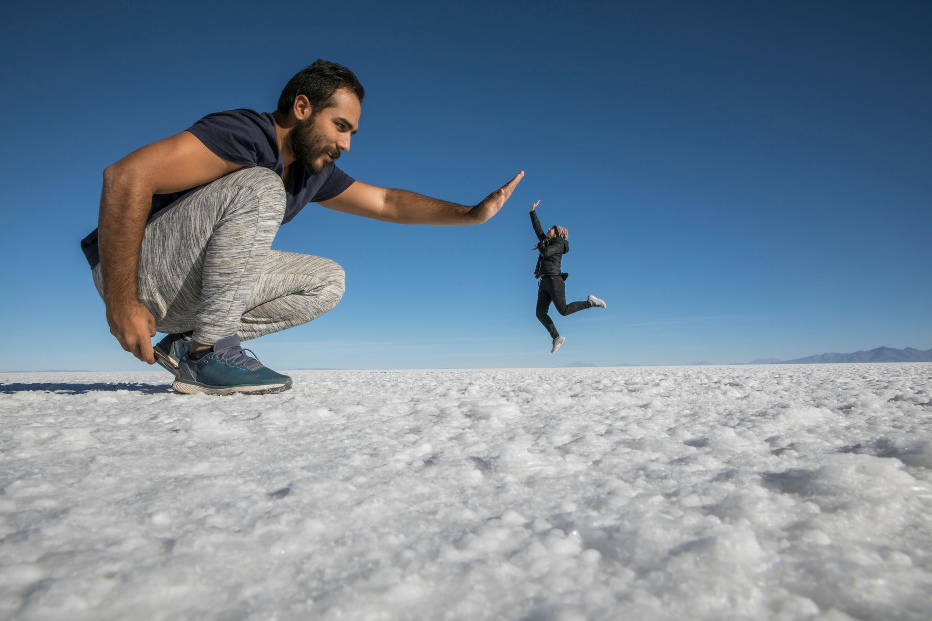 A man is squatting down in the salt flat while a woman is jumping in the air to high five.