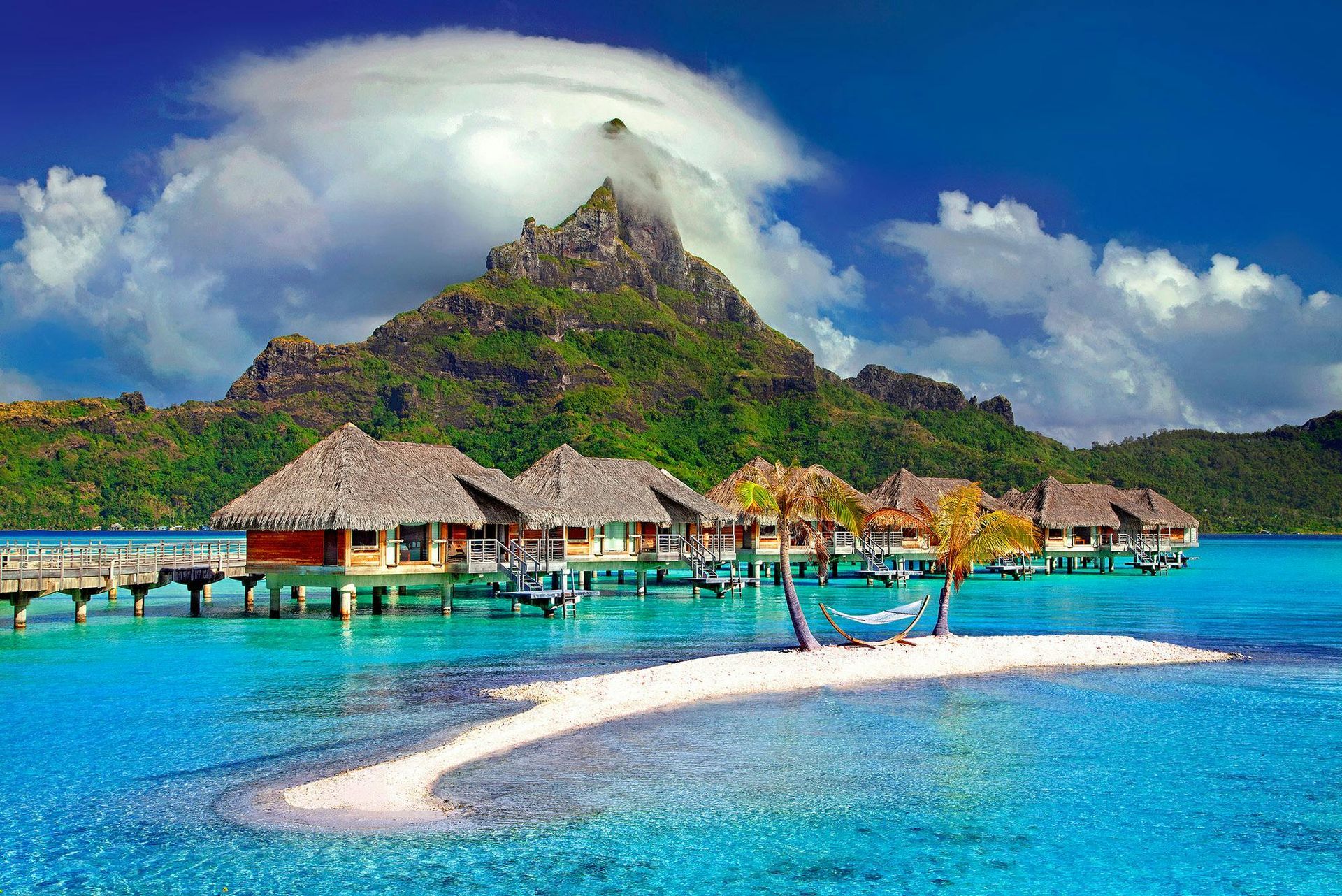 A small island in the middle of the ocean with a mountain in the background with Water bungalows