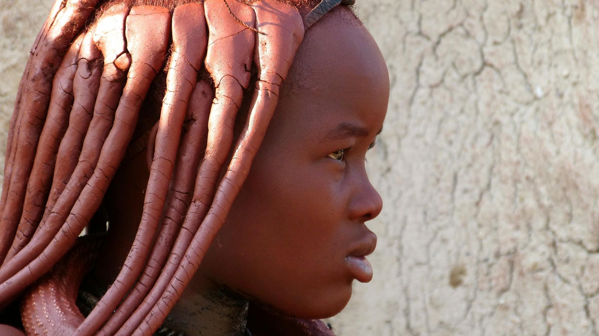 A Himba woman with long red hair is standing in front of a wall in Namibia
