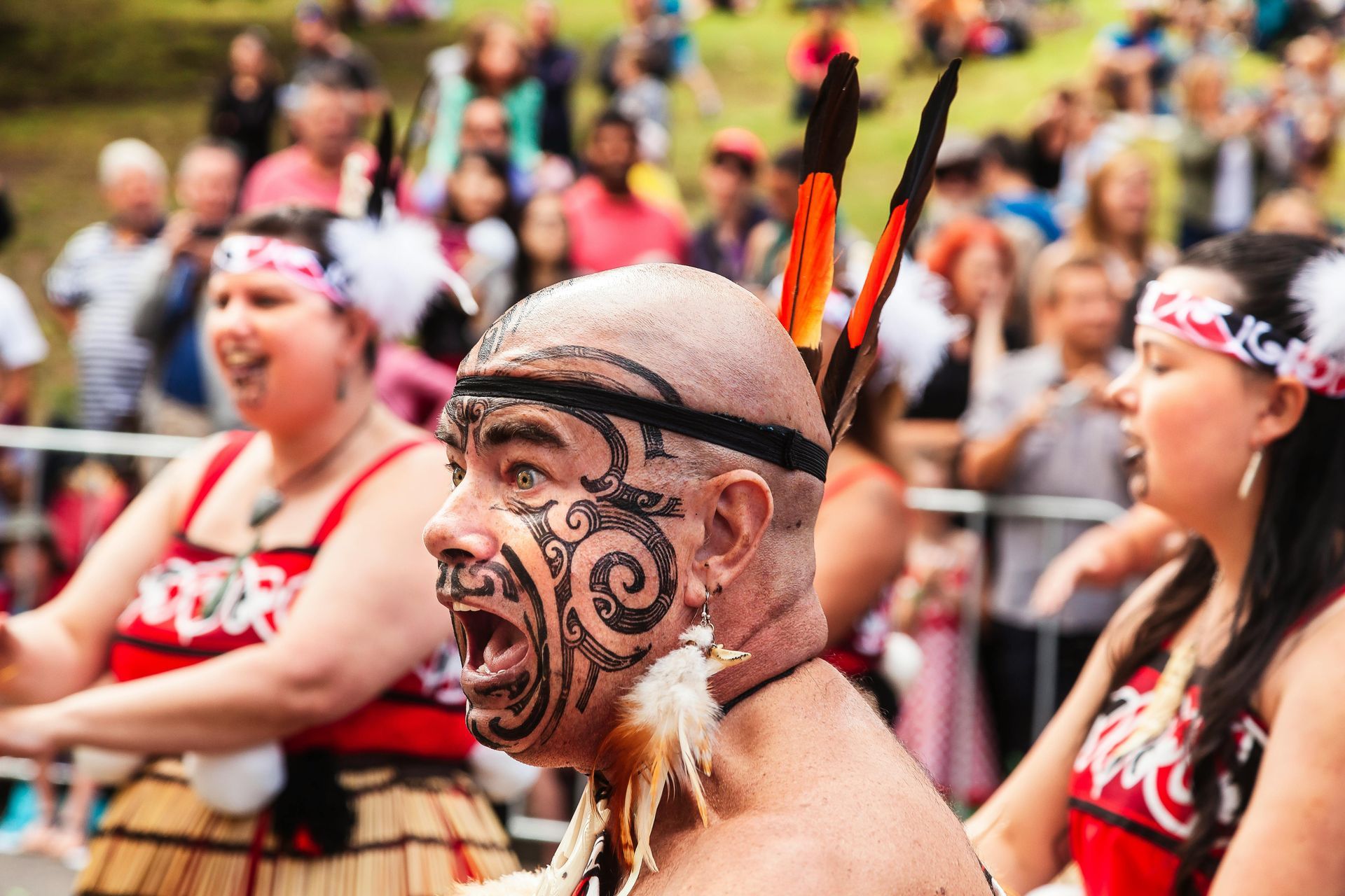 A Maori man with a tattoo on his face is performing a traditional dancing in front of a crowd. 