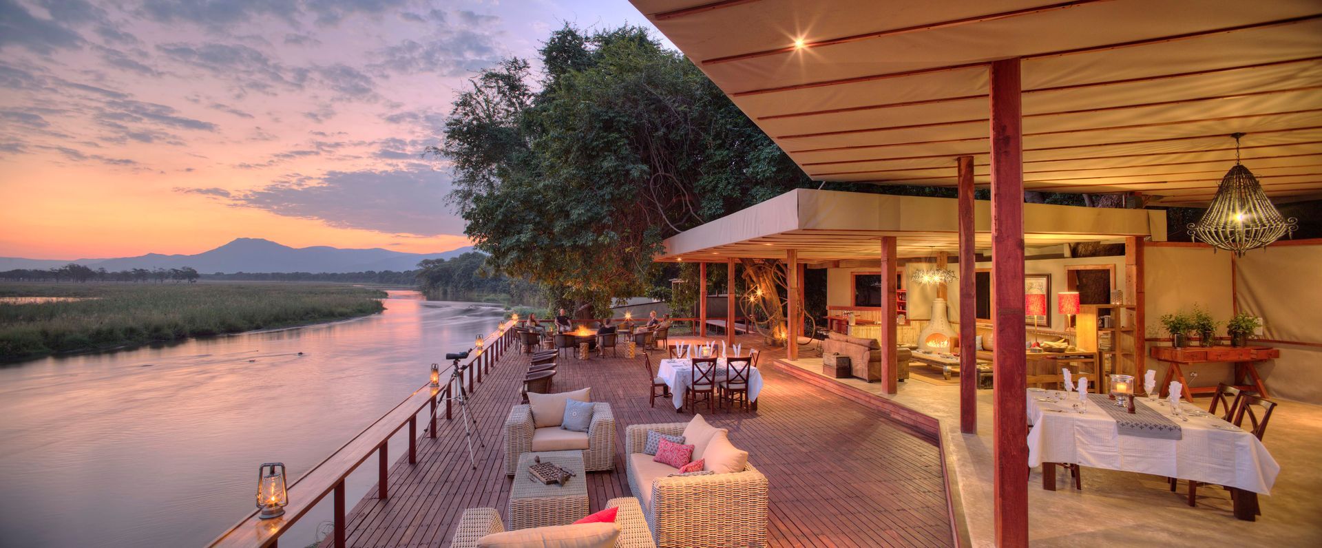 A large patio with tables and chairs overlooking a river at sunset at Sausage Tree Camp