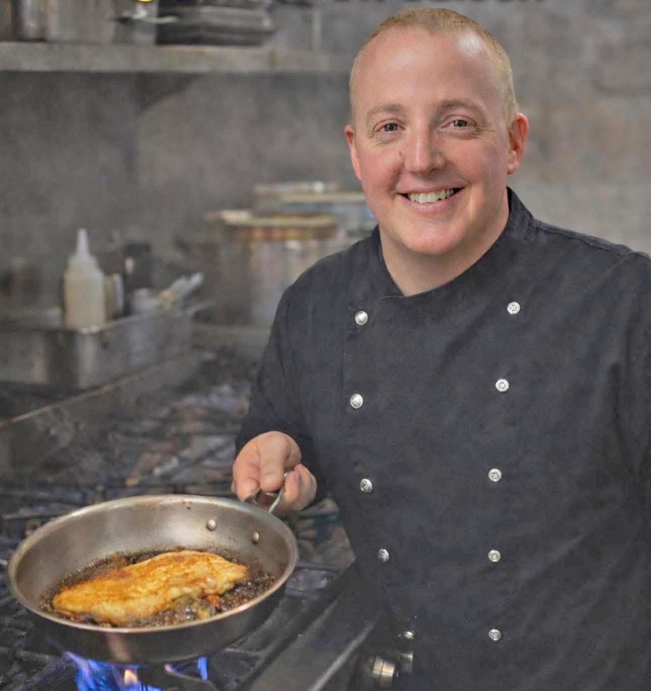 A person in a black chef’s coat leaning against a wooden pillar in a restaurant dining area.