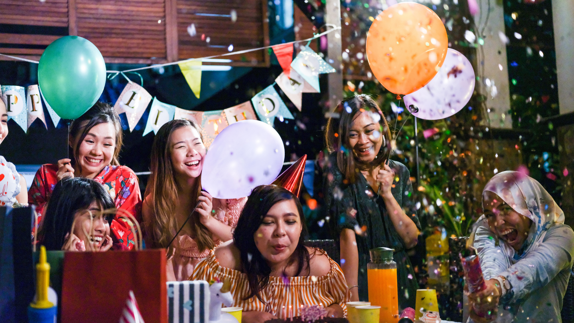 People celebrating a birthday outdoors at night. A woman blows out candles on a cake as confetti and balloons float around.