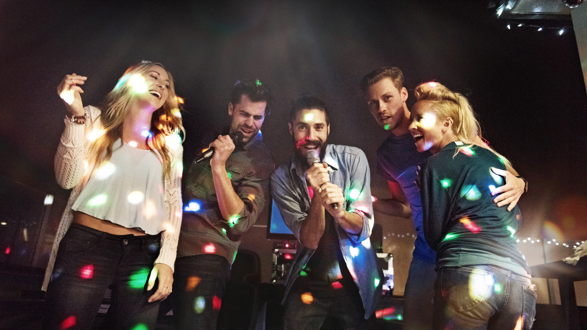 Group of friends enjoying a lively karaoke session, holding microphones and laughing under colorful lights.