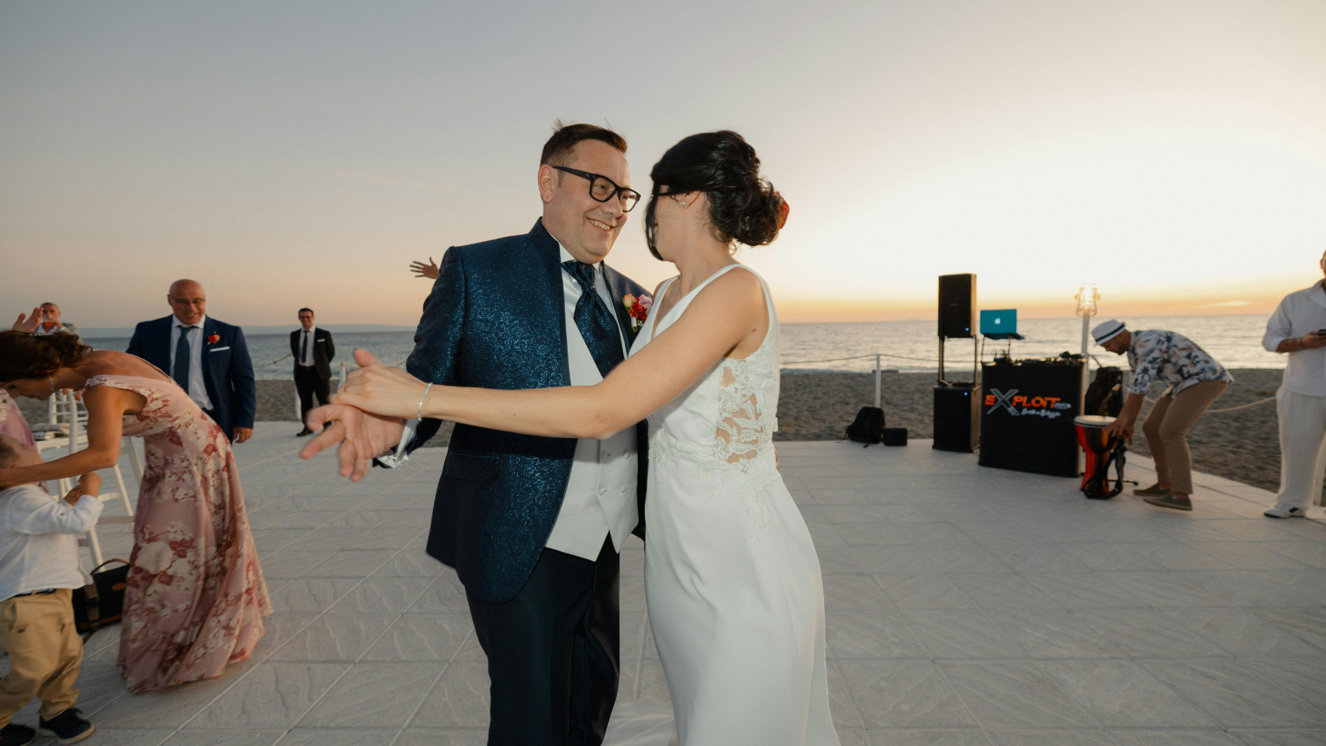 Newlywed couple dancing at a beach wedding with a DJ booth set up in the background and guests enjoying the celebration at sunset.