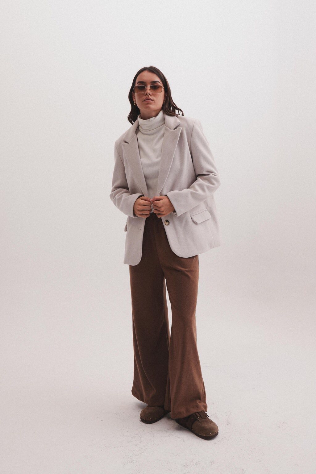 Una mujer que viste una chaqueta blanca y pantalones marrones está parada frente a una pared blanca.