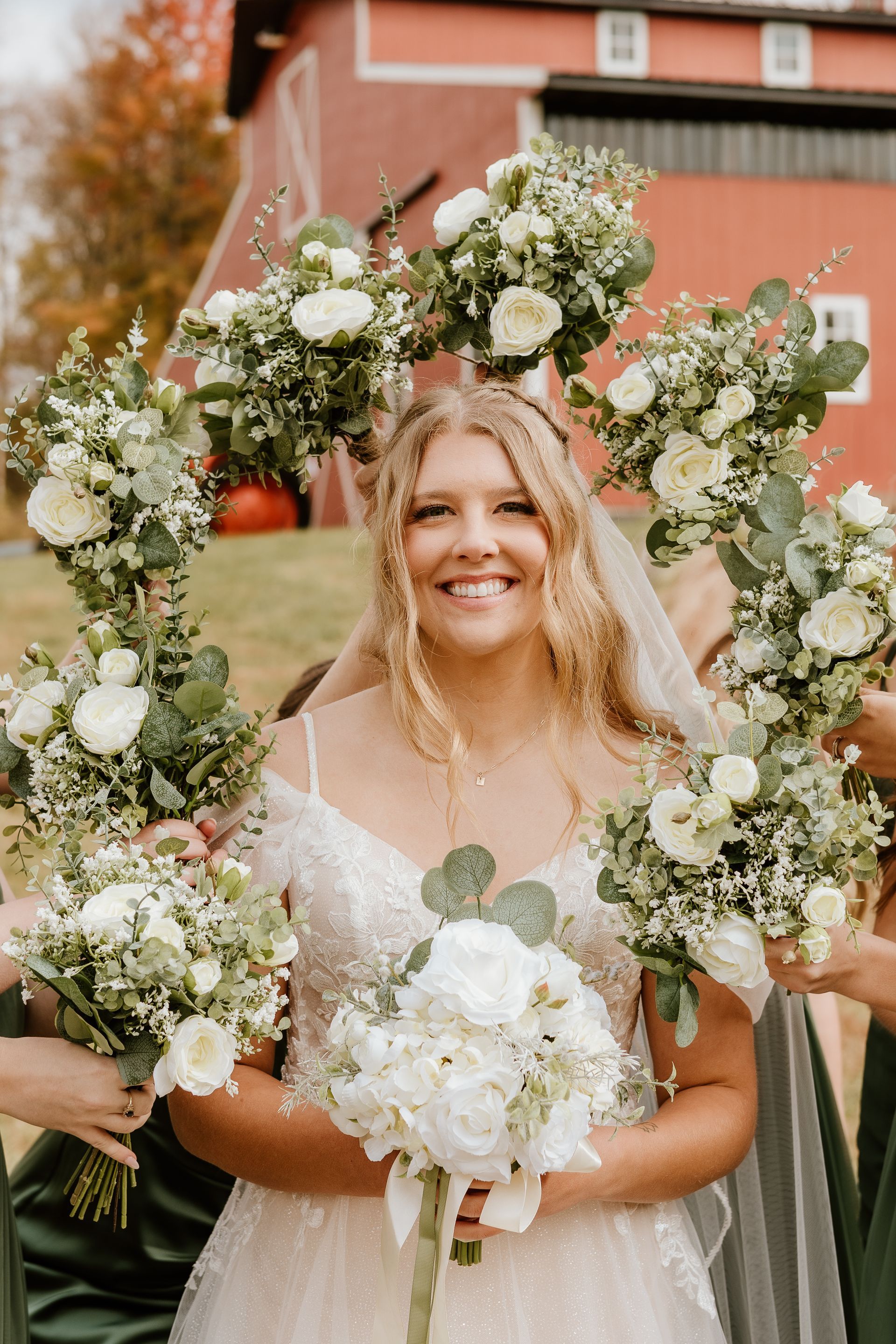 A bride and her bridesmaids are holding bouquets of white flowers in front of a red barn.